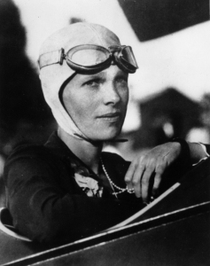 Photographie en noir et blanc d'Amelia Earhart aux commandes d'un avion. Elle porte un bonnet d'aviatrice et des lunettes d'aviatrice remontées sur le front.