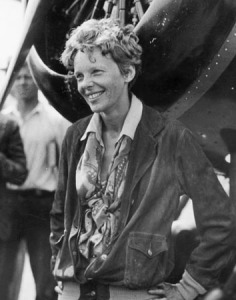 Photographie en noir et blanc d'Amelia Earhart, qui se tient debout, les mains sur les hanches, devant le réacteur d'un avion. Elle sourit.