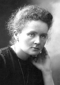 Photographie en noir et blanc de Marie Curie portant un vêtement sombre. La main gauche portée à son visage, elle semble pensive