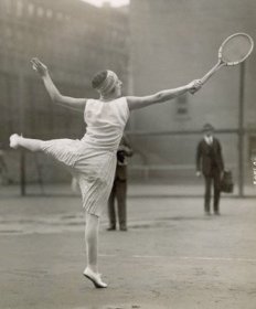 Photographie de Suzanne Lenglen jouant au tennis. Elle tient une raquette au bout de son bras droit tendu et sa jambe gauche est levée toute droite, parallèle au sol