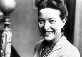 Photographie en noir et blanc de Simone de Beauvoir. Elle porte un collier et de larges boucles d'oreille, et ses cheveux sont retenus en chignon. Elle a un large sourire.