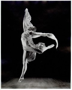 Photographie en noir et blanc d'Isadora Duncan en plein mouvement de danse. Elle saute, une jambe droite et l'autre levée vers l'arrière, les deux bras écartés.