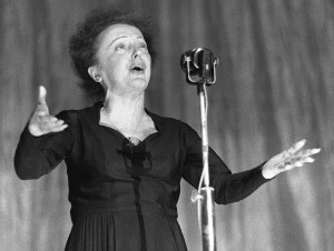 Photographie en noir et blanc d'Edith Piaf en train de chanter, debout devant son micro, les bras écartés devant elle