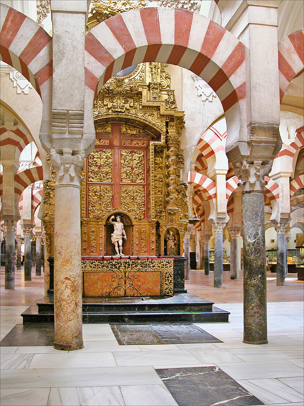 Cette photo représente la Mezquita, la mosquée-cathédrale, de Cordoue. On y voit les colonnades surplombées de pierres rouges et blanches et, au centre de l'image, un autel chrétien.