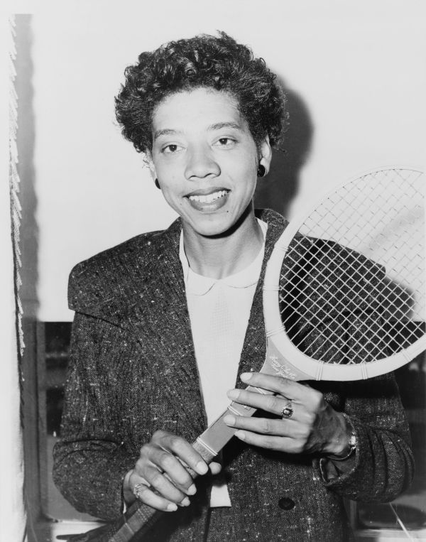 Cette photographie en noir et blanc montre Althea Gibson une raquette de tennis à la main. Elle a les cheveux courts, porte des boucles d'oreille et sourit.