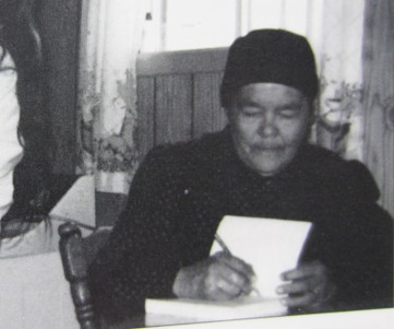 Photographie en noir et blanc d'An Antane Kapesh. Assise à une table, elle porte des habits et un bonnet noir et écrit dans un cahier.