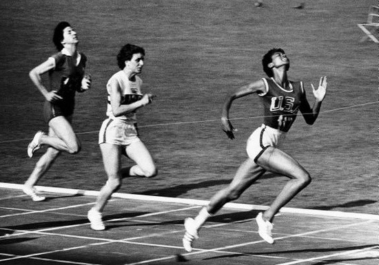 Cette photographie en noir et blanc montre Wilma Rudolph passant la ligne d'arrivée lors des Jeux Olympiques et remportant la médaille d'or. Deux de ses concurrentes sont derrière elle.