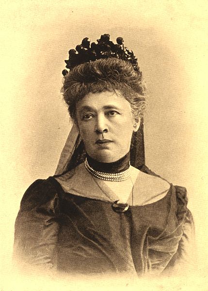 Cette image est un portrait de Bertha von Suttner. Elle porte une riche robe d'époque en tissu sombre, un collier de perles à plusieurs rangs et une coiffure sophistiquée.
