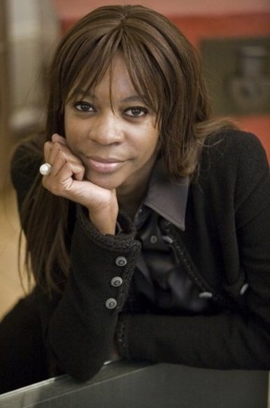 Cette image est une photographie de Dambisa Moyo. L'économiste et autrice est accoudée, le menton reposant dans sa main droite. Elle porte une chemise et une veste noire. Elle sourit.