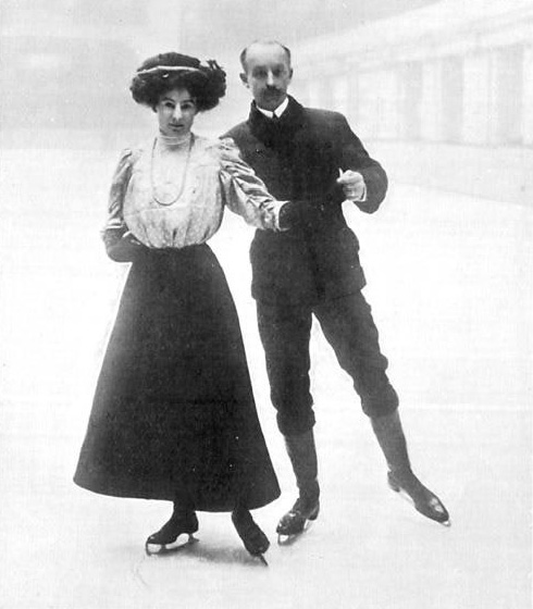 Cette photographie en noir et blanc montre Edgar et Madge Syers aux Jeux olympiques de 1908. Madge, à gauche, porte une jupe sombre comme ses patins et un haut clair. Edgar, à droite, tient Madge par la taille et par la main. Il porte un pantalon et une veste sombres.