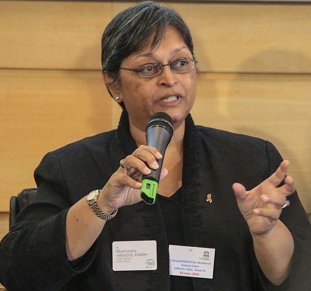 Cette photographie montre Quarraisha Abdool Karim, un micro dans sa main gauche et sa main droite ouverte. Bouche entrouverte, elle est en train d'intervenir lors de la remise des prix L'Oréal-Unesco. Elle a les cheveux courts et grisonnants, porte des lunettes, une veste noire et un montre bracelet.