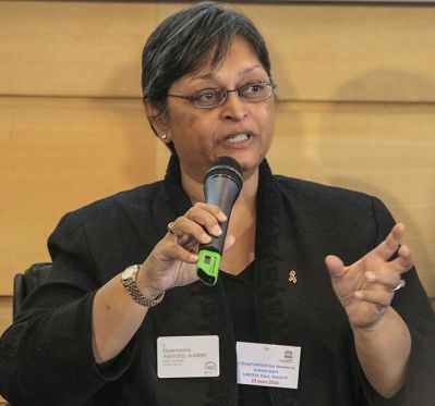 Cette photographie montre Quarraisha Abdool Karim, un micro dans sa main gauche et sa main droite ouverte. Bouche entrouverte, elle est en train d'intervenir lors de la remise des prix L'Oréal-Unesco. Elle a les cheveux courts et grisonnants, porte des lunettes, une veste noire et un montre bracelet.