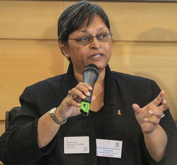 Cette photographie montre Quarraisha Abdool Karim, un micro dans sa main gauche et sa main droite ouverte. Bouche entrouverte, elle est en train d'intervenir lors de la remise des prix L'Oréal-Unesco. Elle a les cheveux courts et grisonnants, porte des lunettes, une veste noire et un montre bracelet.