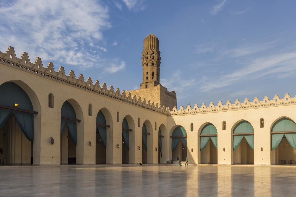 Cette photographie montre la cour de la Mosquée Al-Hakim au Caire, sous un ciel bleu avec quelques nuages de beau temps.