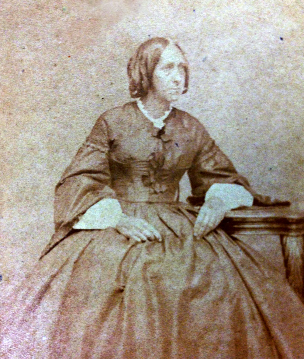 Cete photo ancienne et abimée montre Sarah Harris Fayerweather en robe d'époque sombre, assise sur une chaise et accoudée sur un guéridon.