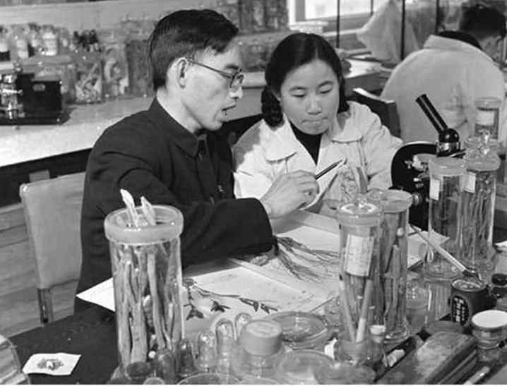 Cette photo en noir et blanc montre Tu Youyou en blouse blanche dans un laboratoire, entourée de matériel scientifique. Elle est assise à côté d'un autre scientifique qui lui montre quelque chose.