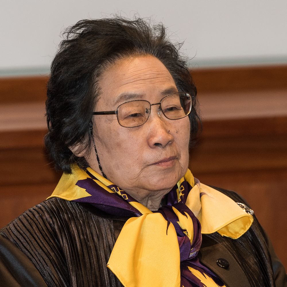 Cette photographie montre Tu Youyou à Stockholm en 2015 lors de la remise de son prix Nobel. Elle porte un foulard jaune et violet, une veste noire et des lunettes.