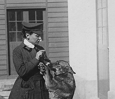 Cette photographie en noir et blanc montre Nicole Girard-Mangin avec sa chienne Dun (pour Verdun). Nicole est débout, en jupe et veste sombre avec une casquette et joue avec sa chienne, debout sur ses pattes arrières.