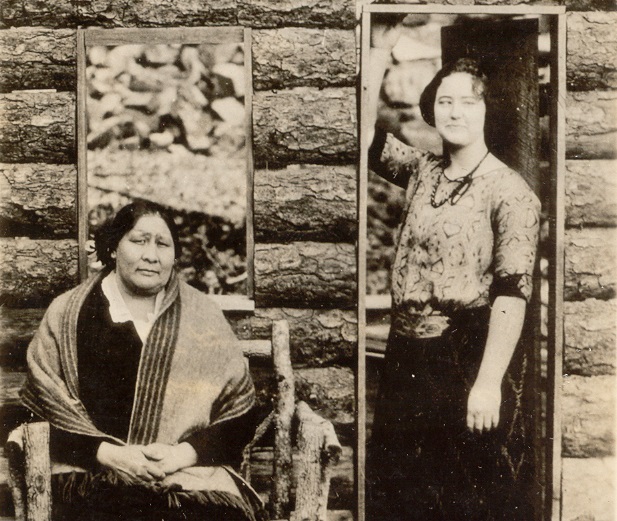 Cette photographie montre Rita Smith et Nettie Brookshire. Les deux femmes se tiennent devant une maison aux murs de bois, l'une est assise et l'autre debout.