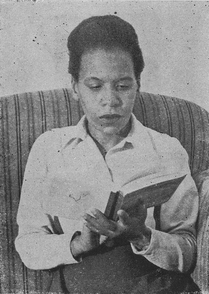 Cette photographie en noir et blanc montre Virginia Brindis de Salas, assise sur un canapé, absorbée dans la lecture d'un livre ouvert entre ses mains. Elle porte une chemise claire.