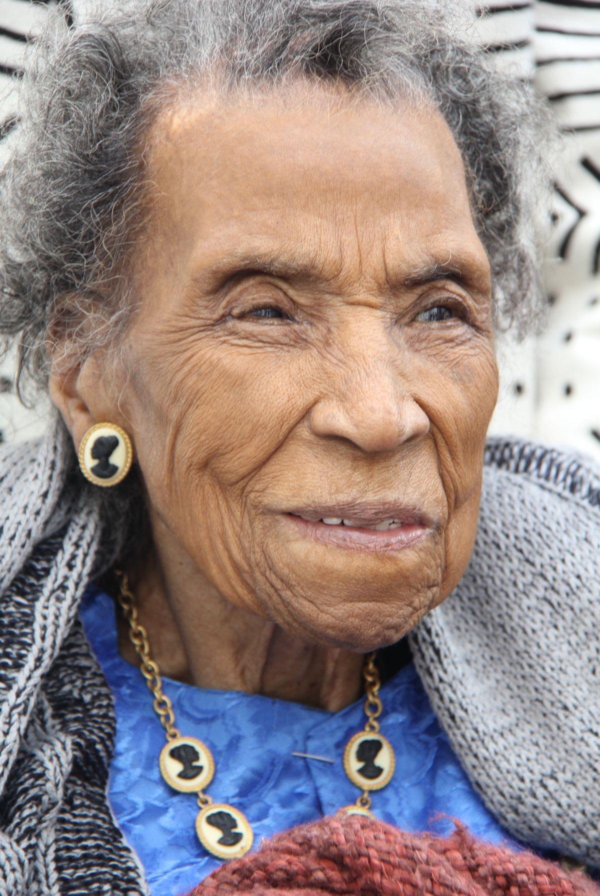 Cette image est un portrait d'Amelia Boyton Robinson, en 2015, à l'âge de 103 ans. Elle a les cheveux gris et un visage souriant. Elle porte une chemise bleue sous un vêtement épais de laine. Elle porte des camées représentant un profil de femme noire en collier et en boucle d'oreille.