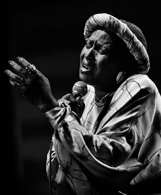 Cette photo en noir et blanc montre Miriam Makeba en train de chanter, micro à la main. Absorbée par sa prestation, elle a les yeux fermés, les lèvres ouvertes et la main gauche levée, tandis que l'autre tient le micro.