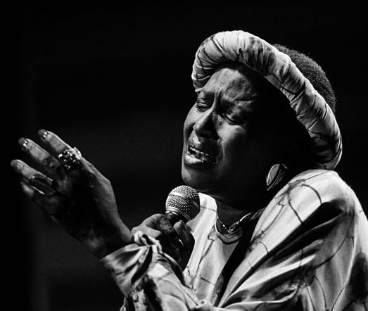 Cette photo en noir et blanc montre Miriam Makeba en train de chanter, micro à la main. Absorbée par sa prestation, elle a les yeux fermés, les lèvres ouvertes et la main gauche levée, tandis que l'autre tient le micro.