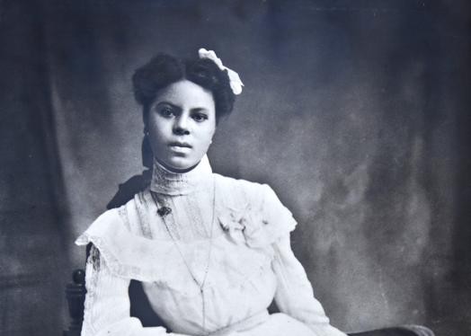 Cette photographie en noir et blanc montre Lucile Buchanan, assise sur une chaise en bois et vêtue d'une robe blanche sophistiquée avec des manches longues. Le bras droit appuyé sur un accoudoir de la chaise, elle regarde directement la caméra.
