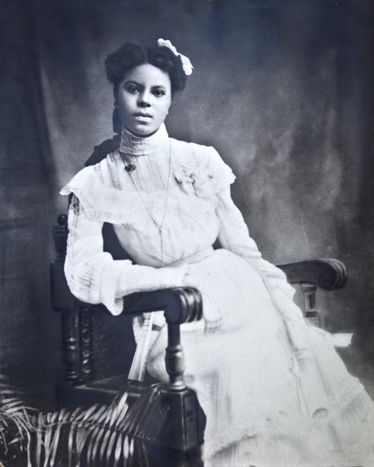 Cette photographie en noir et blanc montre Lucile Buchanan, assise sur une chaise en bois et vêtue d'une robe blanche sophistiquée avec des manches longues. Le bras droit appuyé sur un accoudoir de la chaise, elle regarde directement la caméra.