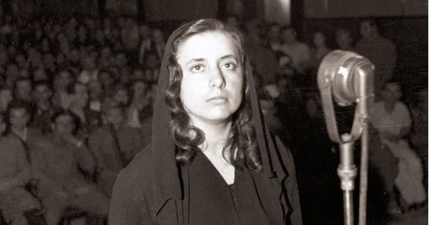 Cette photographie en noir montre Musine Kokalari lors de son procès en 1946. Au premier plan, elle se tient debout devant un micro, vêtue d'habits sombres et un voile couvrant ses cheveux. Elle a l'air grave. Derrière elle, à l'arrière plan, des rangs de spectateurs assis et debout assistent au procès.