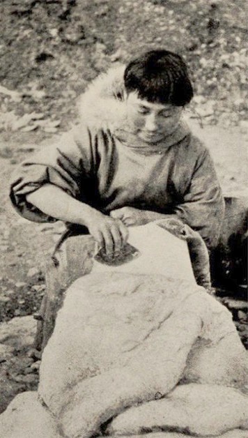 Cette photographie en noir et blanc montre Ada Blackjack assise ; elle est occupée à enlever la graisse d'une peau de phoque.