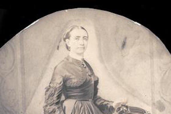 Cette image est une aquarelle en noir et blanc représentant Kate Warne debout devant un fauteuil, vêtue d'une robe sombre et longue à la jupe large, la main gauche posée sur le fauteuil. Elle a les cheveux tirés en arrière.