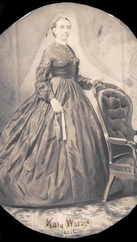 Cette image est une aquarelle en noir et blanc représentant Kate Warne debout devant un fauteuil, vêtue d'une robe sombre et longue à la jupe large, la main gauche posée sur le fauteuil. Elle a les cheveux tirés en arrière.