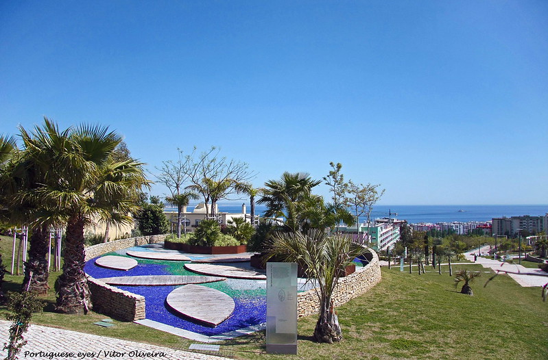 Cette photographie montre un monument à Alda do Espirito Santo dans un parc au Portugal. Au premier plan, le monument avec des bassins et des palmiers, à l'arrière-plan la mer.