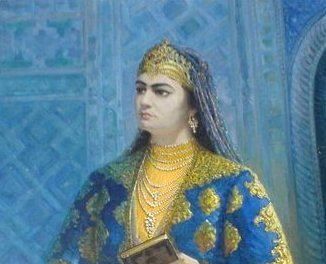 Cette illustration montre Nadira, vêtue d'une robe bleue et jaune. Elle porte une couronne, un collier avec de nombreux rangs de perles, des bracelets et des bagues. Un voile recouvre ses cheveux. Elle tient un livre entre ses deux mains.