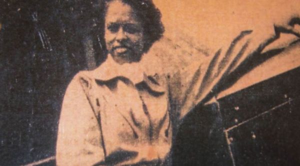 Cette image est une vieille photographie montrant Azellia White devant une avion, la main gauche posée sur l'avion. Elle sourit.