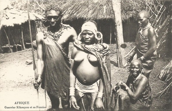 Cette carte postale en noir et blanc du début du 20e siècle montre quatre personnes. A gauche, debout, un guerrier kikuyu accompagné de sa femme ; à droite, une autre personne assise et une debout. En arrière-plan, on distingue deux cases.