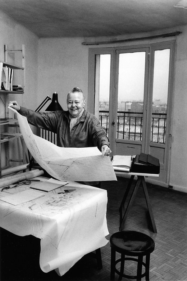 Cette photo en noir et blanc de Robert Doisneau montre Charlotte Perriand dans son atelier (à l'age de 78 ans). Installée derrière un bureau couvert de papiers et de crayons, elle tient un grand schéma des deux mains. Elle est vêtue d'une chemise, prote ses cheveux en chignon et affiche un large sourire. Derrière elle, une fenêtre permet de voir un environnement urbain.