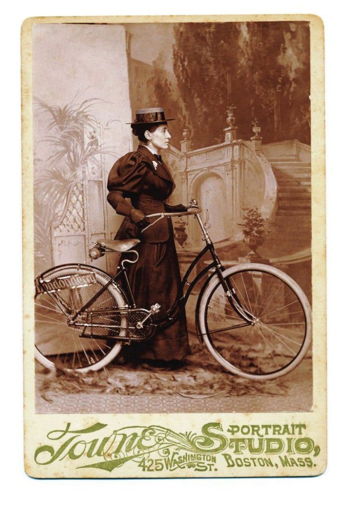 Cette photographie montre Annie Londonderry debout, vêtue d'une longue robe sombre et d'un chapeau à ruban, tenant par le guidon un vélo. Elle est devant un arrière-plan montrant un escalier monumental et des arbres.