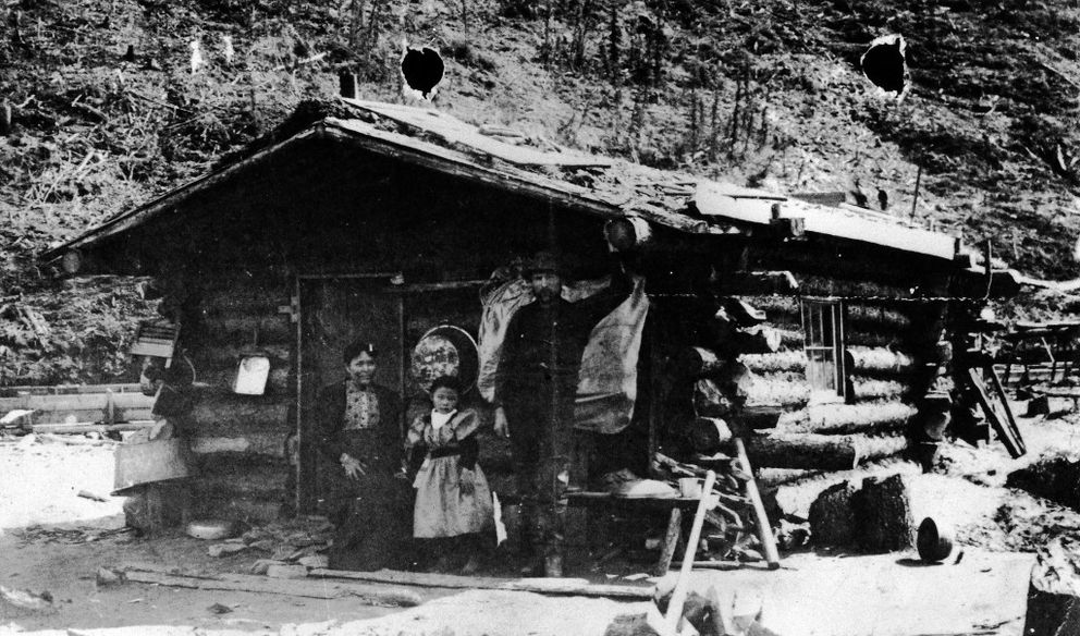 Cette photographie en noir et blanc montre Kate Carmack, Graphie Carmack et George Karmak devant leur cabane de bois. Kate est assise devant la porte tandis que sa fille et son mari se tiennent debout.