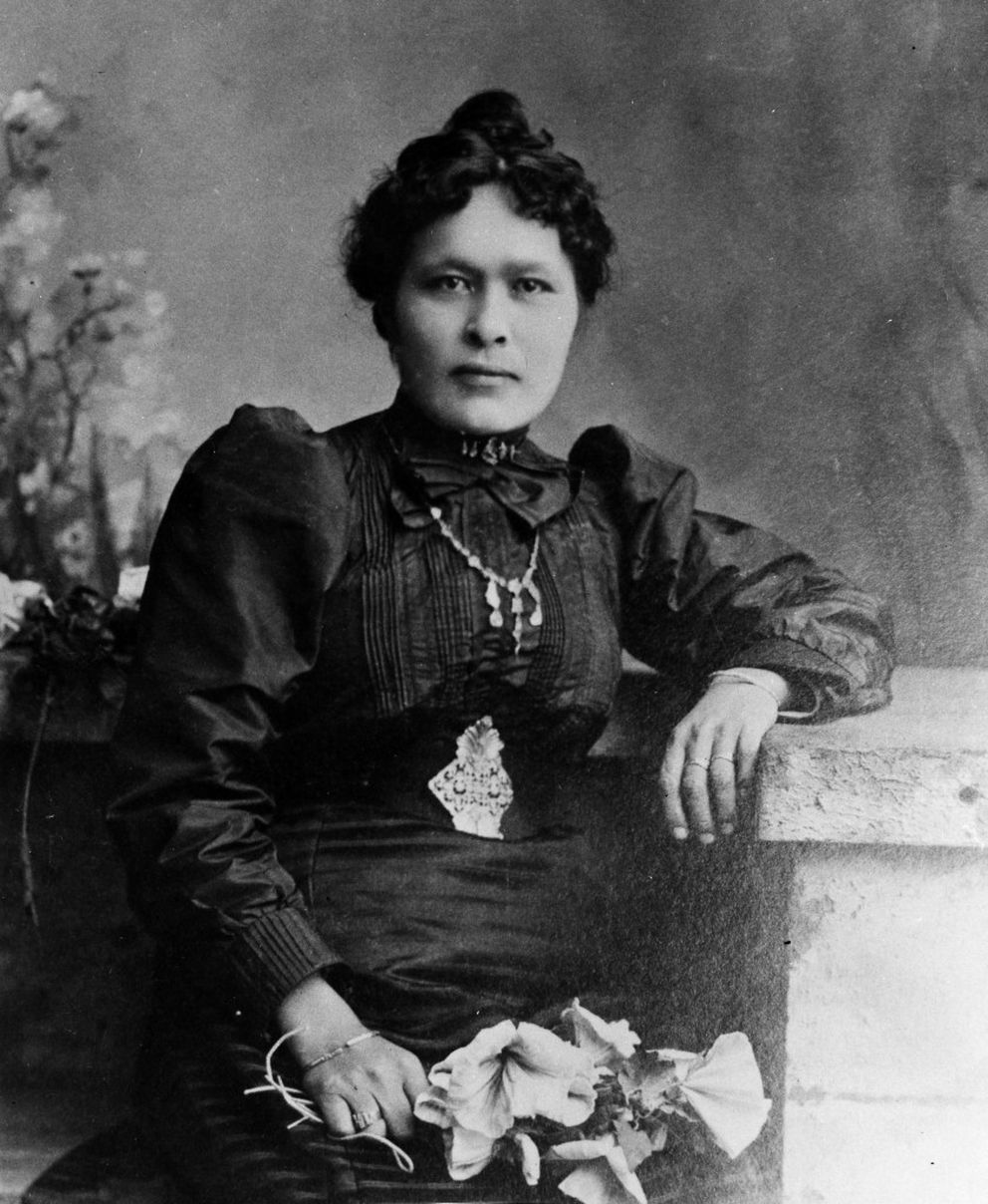 Cette photographie en noir et blanc  montre  Shaaw Tláa - Kate Carmack en riches vêtements occidentaux. Elle porte une longue robe sombre, une ceinture et un collier ouvragés et tient un bouquet de fleurs à la main.