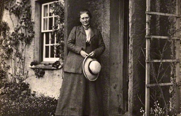 Cette image en noir en blanc montre Beatrix Potter devant sa maison. Vêtue d'une longue jupe sombre et d'une veste assortie, elle tient un chapeau de paille entre ses mains.