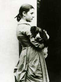 Cette photographie en noir et blanc montre Beatrix Potter à l'âge de 15 ans. De profil, vêtue d'un long manteau, les cheveux retenus en chignon, elle tient un chien dans ses bras.