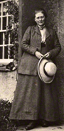 Cette photographie en noir et blanc montre Beatrix Potter, debout devant sa maison. Elle est vêtue d'une jupe longue et d'une veste et tient un chapeau entre ses mains.