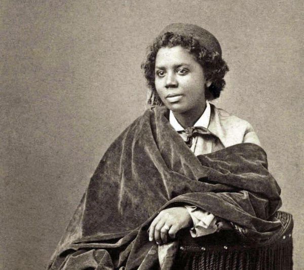 Cette image est une photographie en noir et blanc d'Edmonia Lewis. Elle est assise sur un fauteuil et porte une longue robe. Son buste est couvert d'un grand châle sombre.