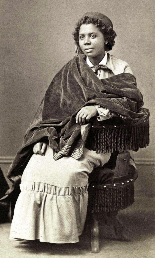Cette image est une photographie en noir et blanc d'Edmonia Lewis. Elle est assise sur un fauteuil et porte une longue robe. Son buste est couvert d'un grand châle sombre.