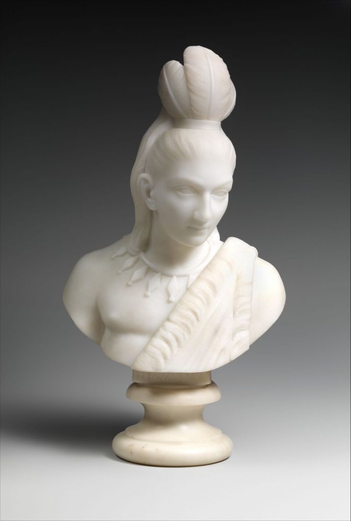 Cette image montre l'oeuvre Hiawatha d'Edmonia Lewis ; il s'agit d'un portrait en buste de marbre d'une personne amérindienne portant une coiffe, un collier et un vêtement traditionnels.