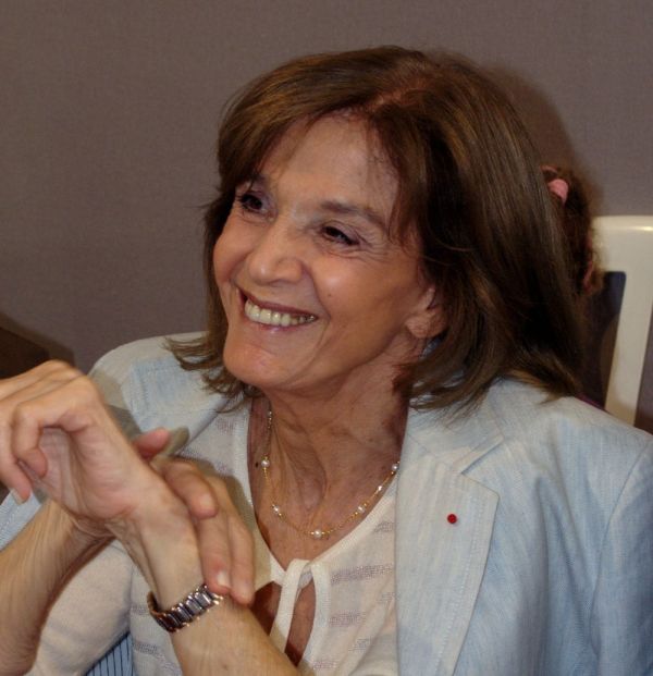 Cette photographie est un portrait de Gisèle Halimi en 2008. Elle est assise, porte un haut blanc et une veste bleue sur les épaules. Ses coudes sont posés sur une table devant elle. Elle sourit.