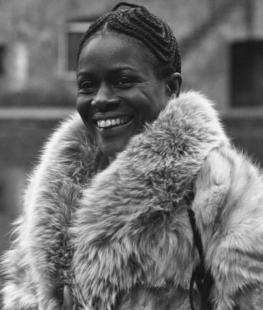 Cette image est un portrait en noir et blanc de Cicely Tyson en 1973. Elle porte un manteau. Ses cheveux sont noués en tresses près du crâne. Elle a un large sourire.
