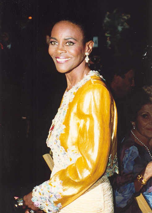 Cette photo est un portrait de Cicely Tyson en 1992. Elle porte une veste jaune brodée aux manches et aux contours, et des boucles d'oreille. Elle a un grand sourire.